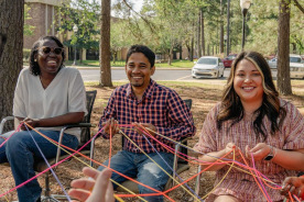 Students and faculty sit in a circle outdoors, working on a team-building exercise with colorful yarn