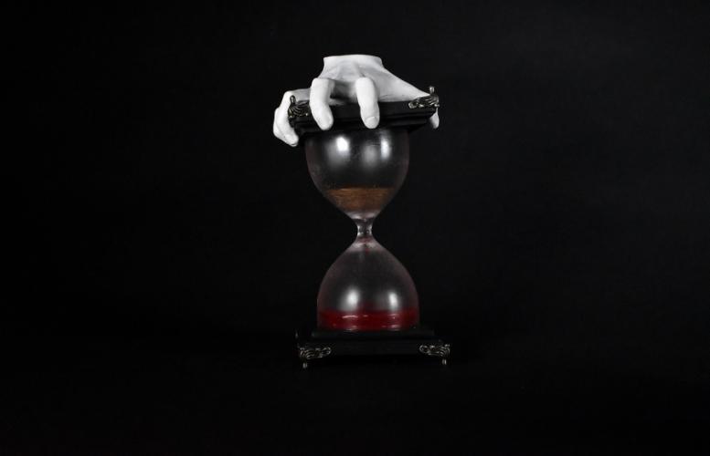 the black and white image of a hand holding an in-color hourglass with what appears to be sand falling down into the bottom of the glass where it looks like the sand has turned to blood