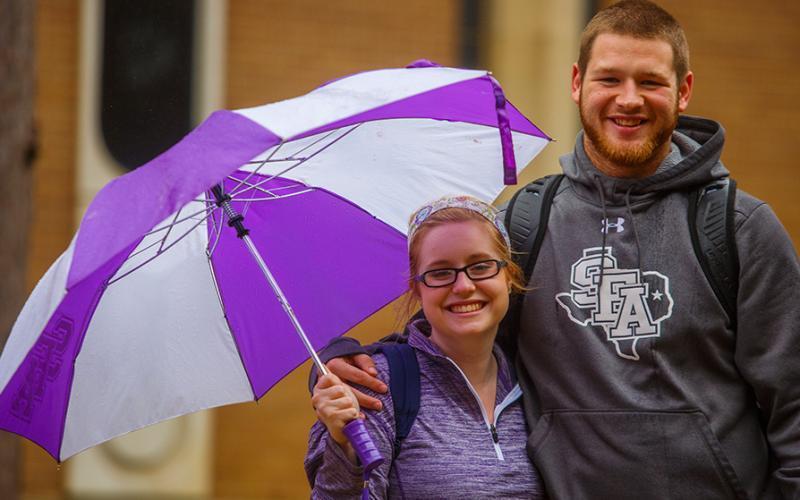 Two students wearing SFA attire stand underneath a purple and white umbrella
