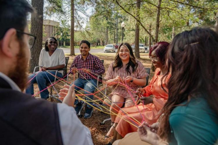 Group of people sitting in a circle outdoors, smiling and holding colorful strings