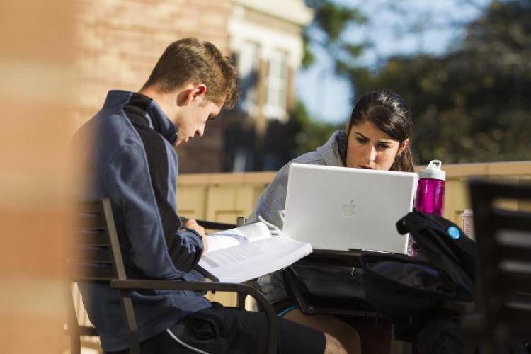 Two students studying together outdoors with a laptop and books
