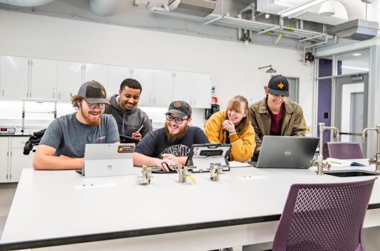 Five students collaborating and smiling while working on laptops in a lab