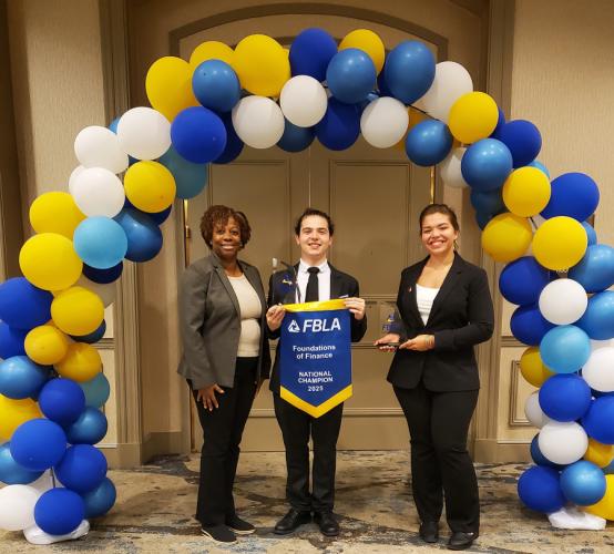 Dr. Marice Kelly and students Julian Ieva and Jessica Frasier stand underneath a balloon arch, holding an FBLA tapestry